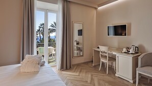 A hotel room with a bed, desk, chairs, a TV, and a balcony with a view of palm trees. - Hotel Paradiso (Sanremo)
