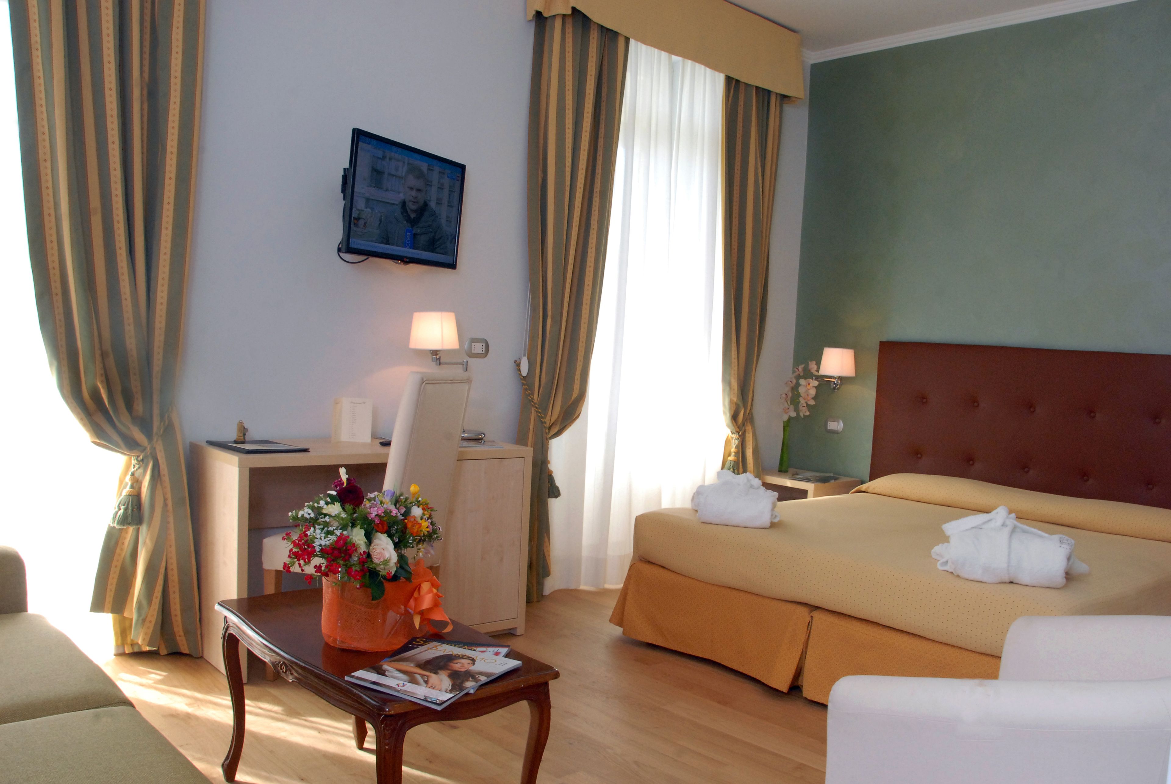 Una habitación de hotel con cama, escritorio, silla, una mesa con un jarrón de flores y televisor en la pared.