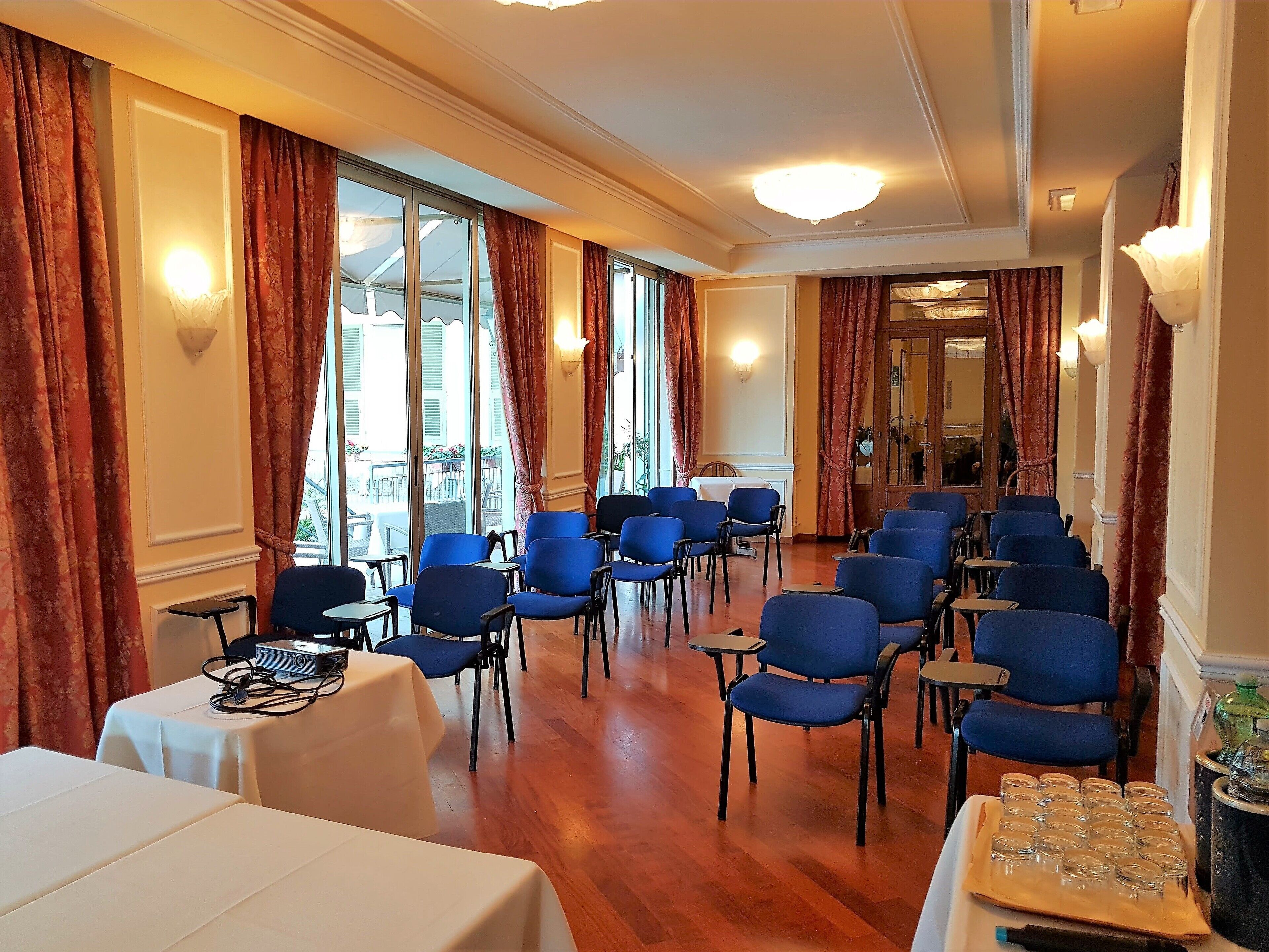 Una habitación de hotel con una mesa larga, varias sillas azules y un ventanal con cortinas.