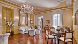 A spacious room with a wooden floor, a chandelier, and a dining area with wooden chairs and a table. - Hotel Paradiso (Sanremo)