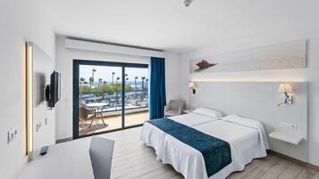 A modern hotel room with a large bed, a desk, and a view of palm trees and a cityscape through a sliding door.
