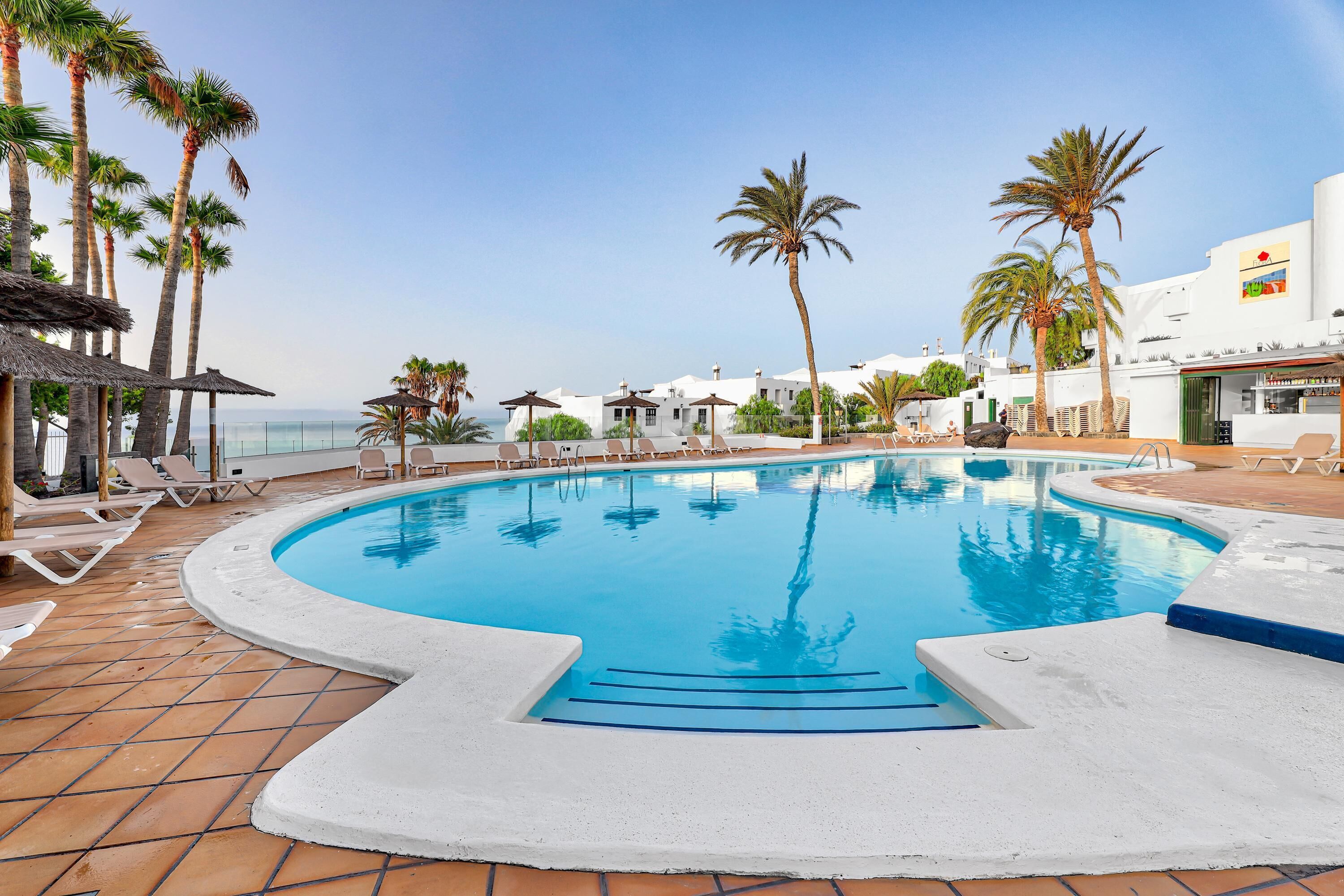 Zona de piscina con vista despejada al océano, rodeada de palmeras y sillas de descanso.