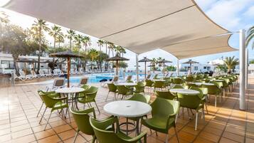 Zona junto a la piscina con sillas y mesas verdes bajo grandes sombrillas, piscina y palmeras al fondo.