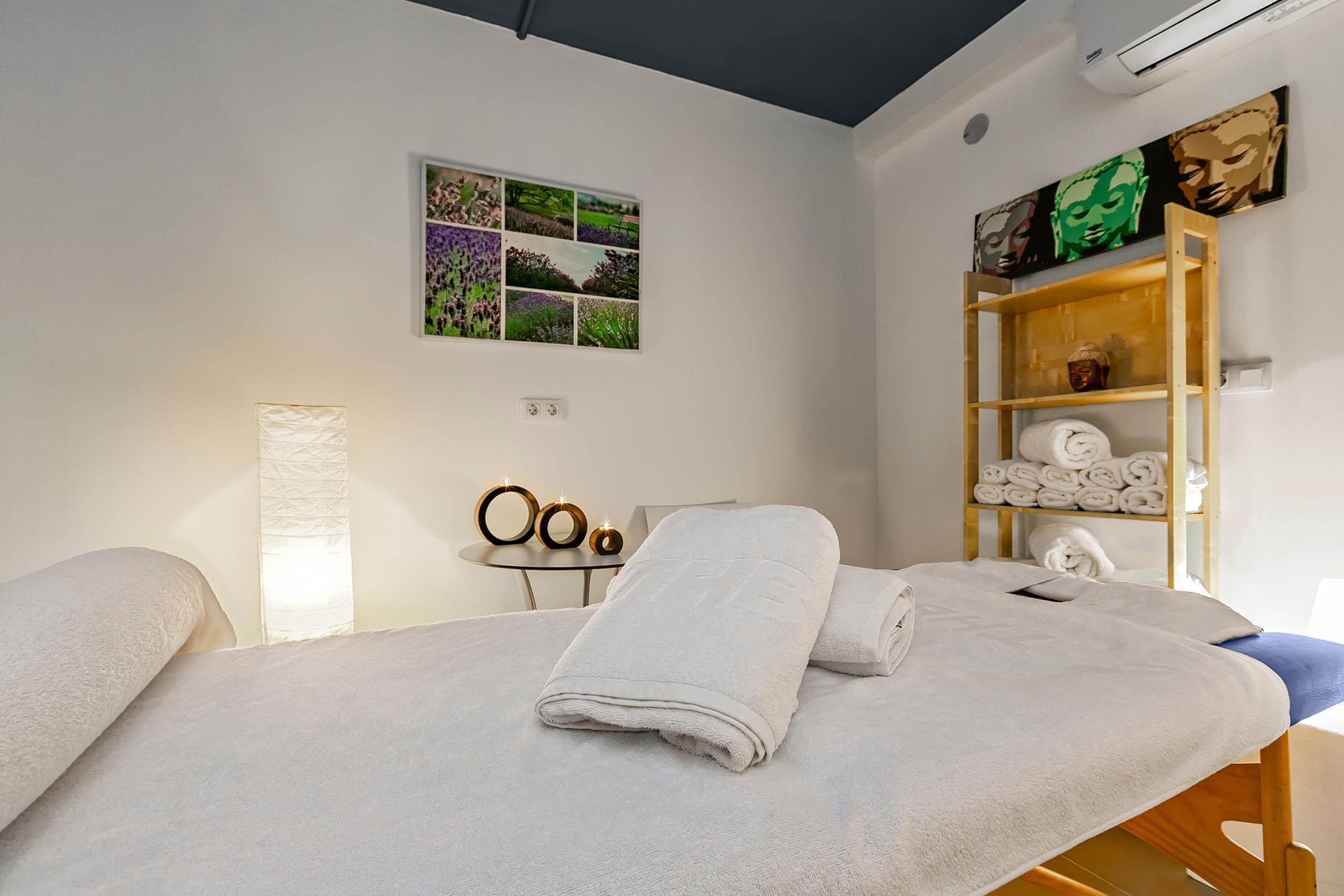 Una habitación con dos camas, un estante con toallas, un collage de fotos enmarcadas y una lámpara.