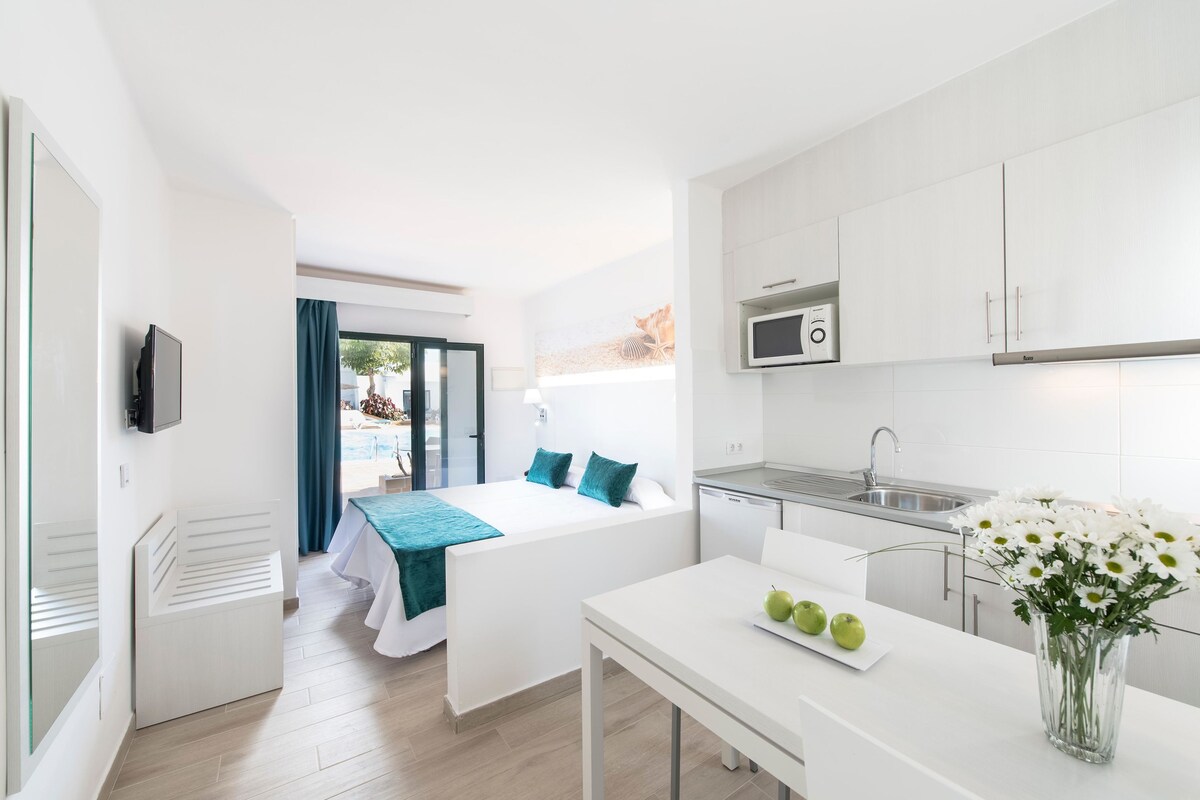 A modern hotel room with a bed, a small desk, a kitchen area with a microwave, and a view of the outdoors.