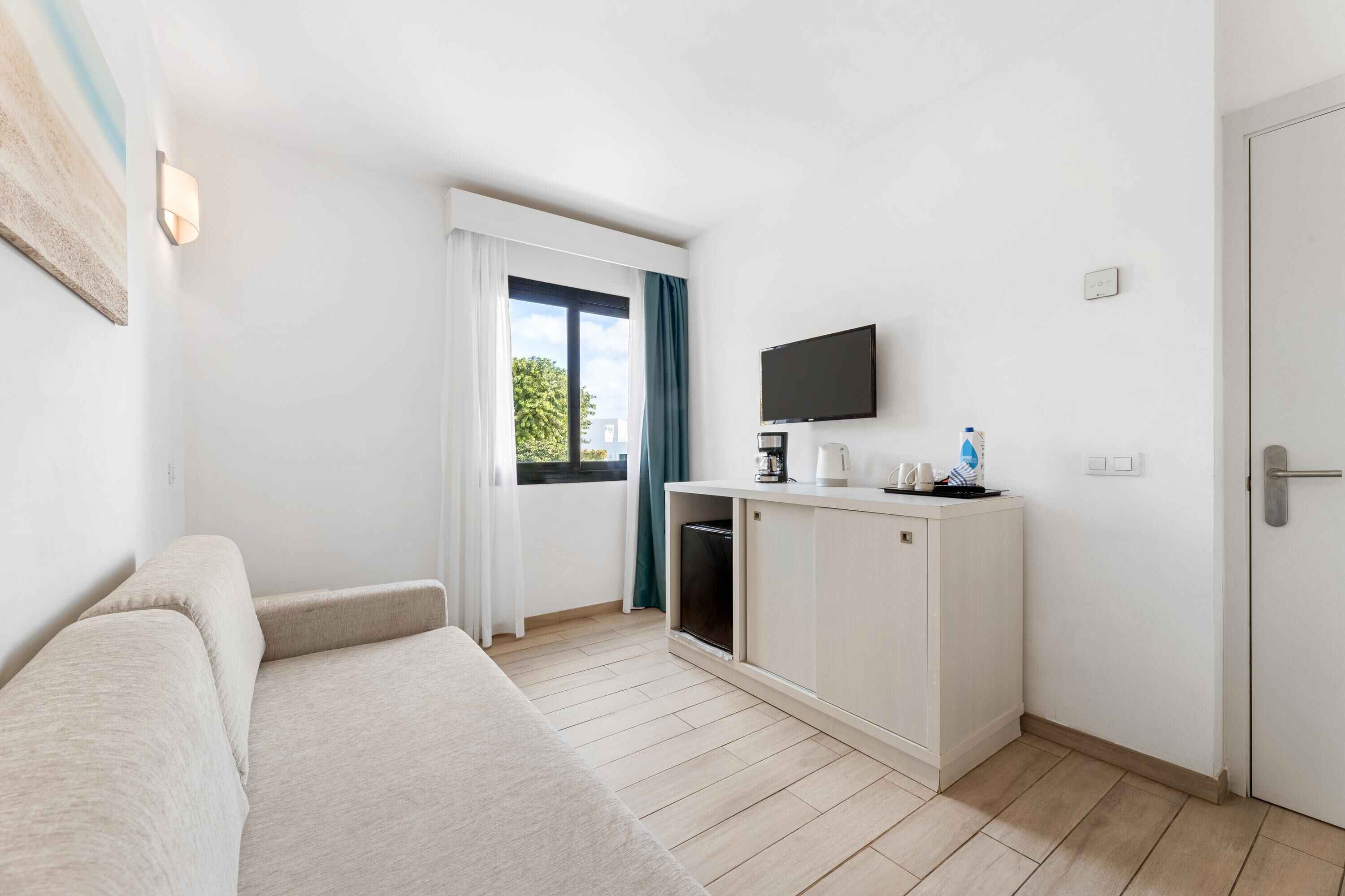 A modern hotel room with a sofa, a small kitchenette, a flat-screen TV, and a sliding glass door to the outside.