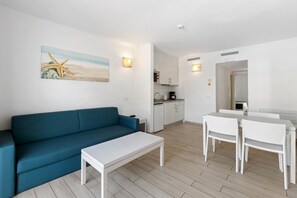 A modern living room with a blue sofa, white coffee table, and dining area with white chairs. There is a kitchen in the background with white cabinets and a coffee machine.
