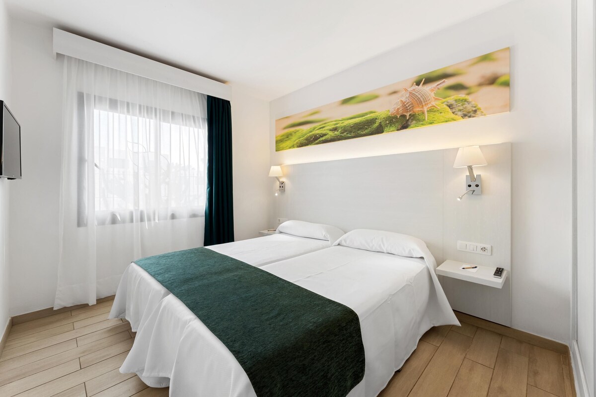 A hotel room with two beds, a large window with sheer curtains, a wall-mounted TV, and a painting above the bed.