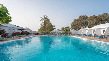 Piscina con sillas de descanso y sombrillas, rodeada de edificios blancos y árboles.