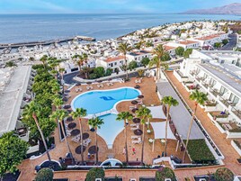 Um resort com piscina central rodeada por espreguiçadeiras e guarda-sóis, edifícios brancos e vista para o mar.