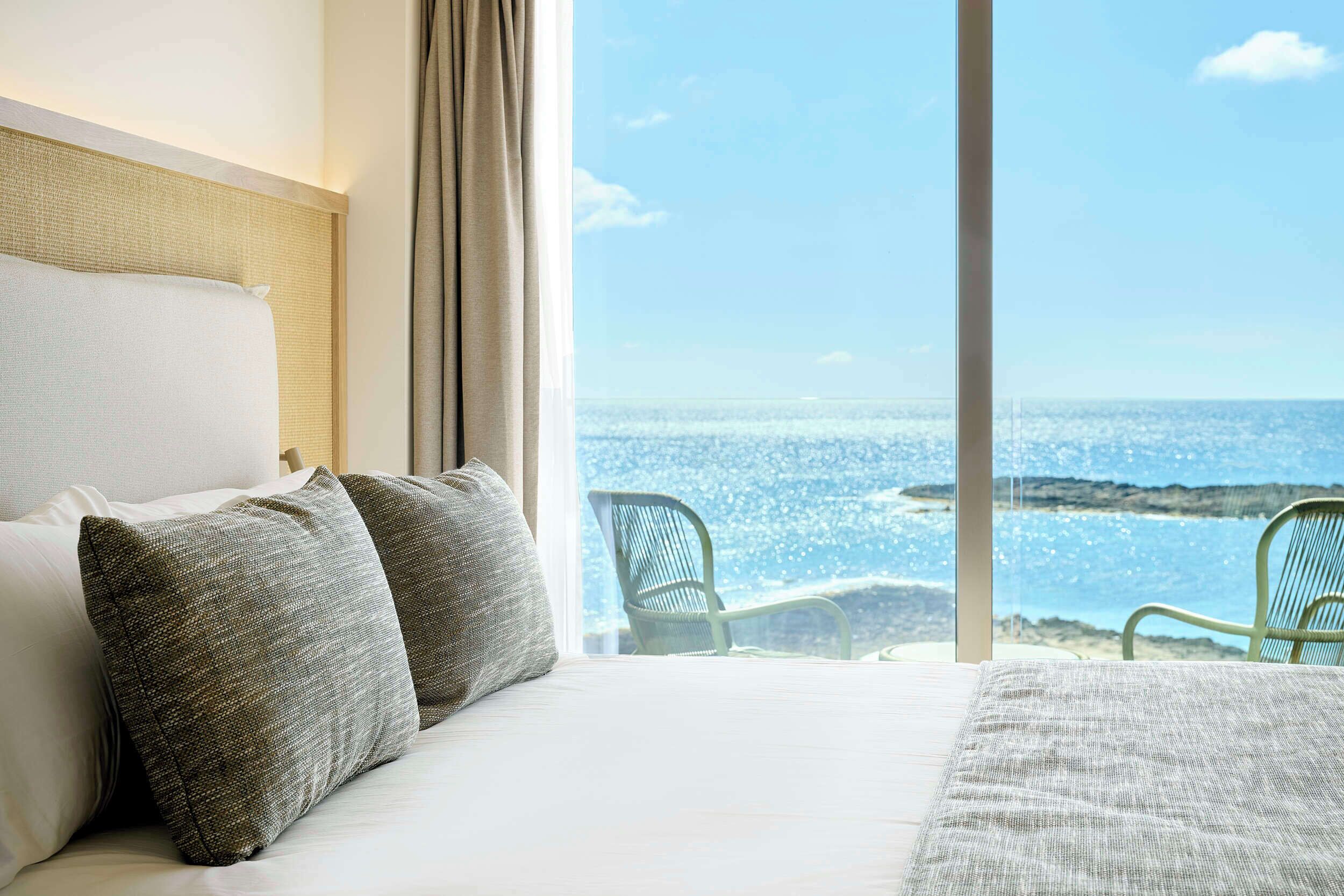 Habitación de hotel con una cama grande, dos almohadas, vista al mar y una silla junto a la ventana.