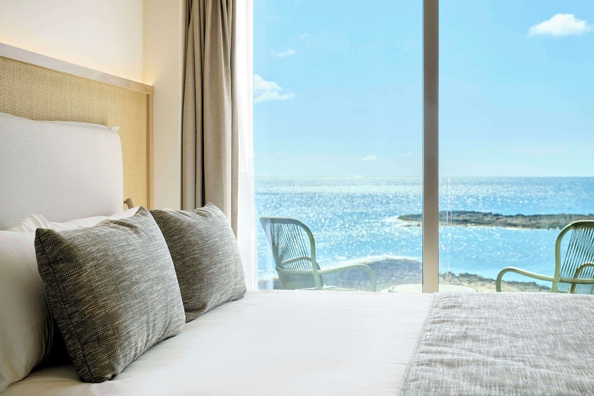 Habitación de hotel con una cama grande, dos almohadas, vista al mar y una silla junto a la ventana.