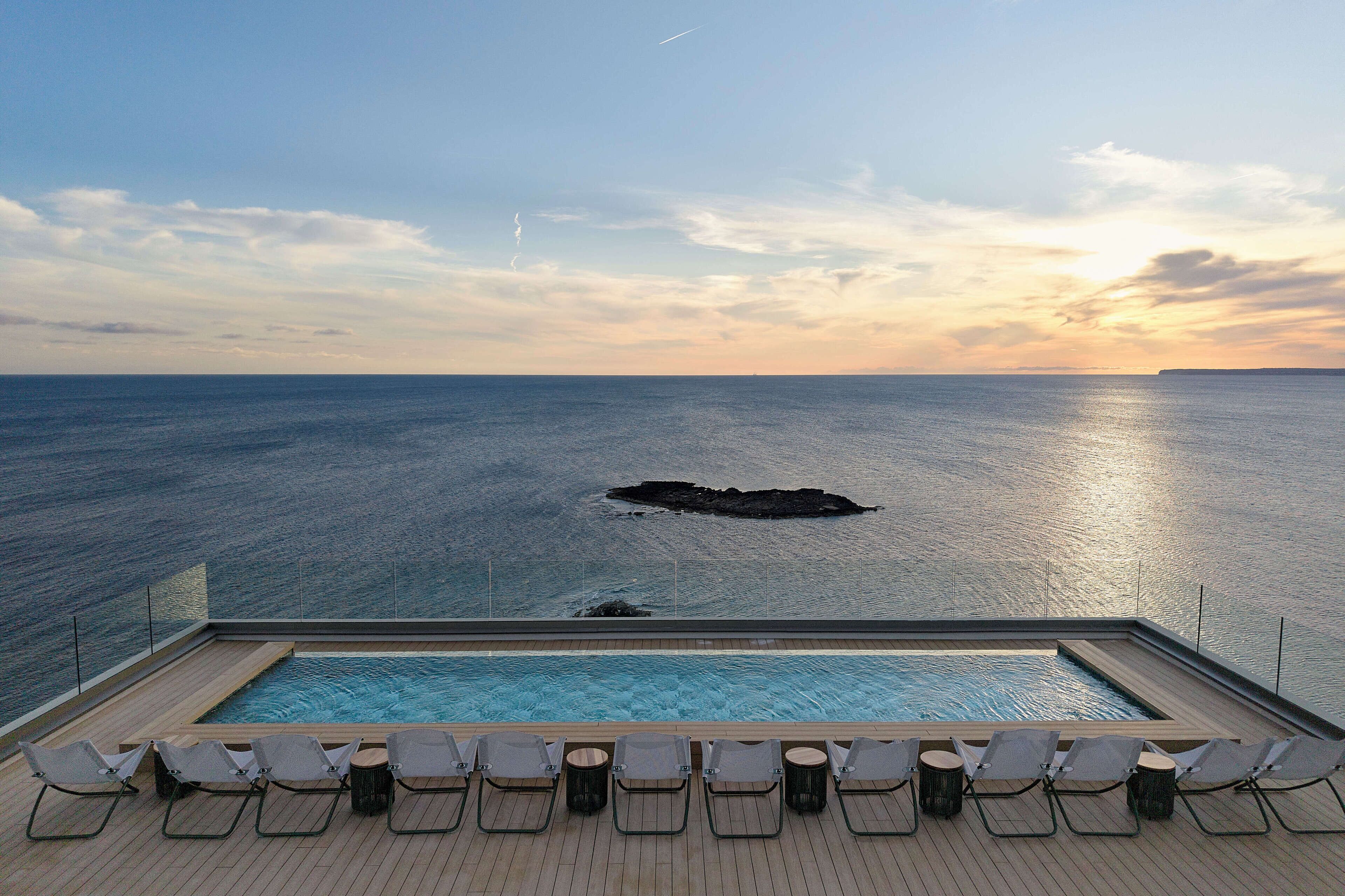 An infinity pool with lounge chairs facing the ocean, a clear sky, and a rocky outcrop in the distance.