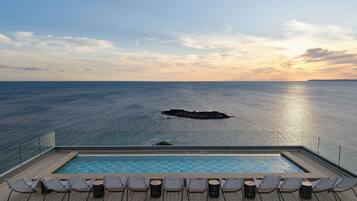 An infinity pool with lounge chairs facing the ocean, a clear sky, and a rocky outcrop in the distance.