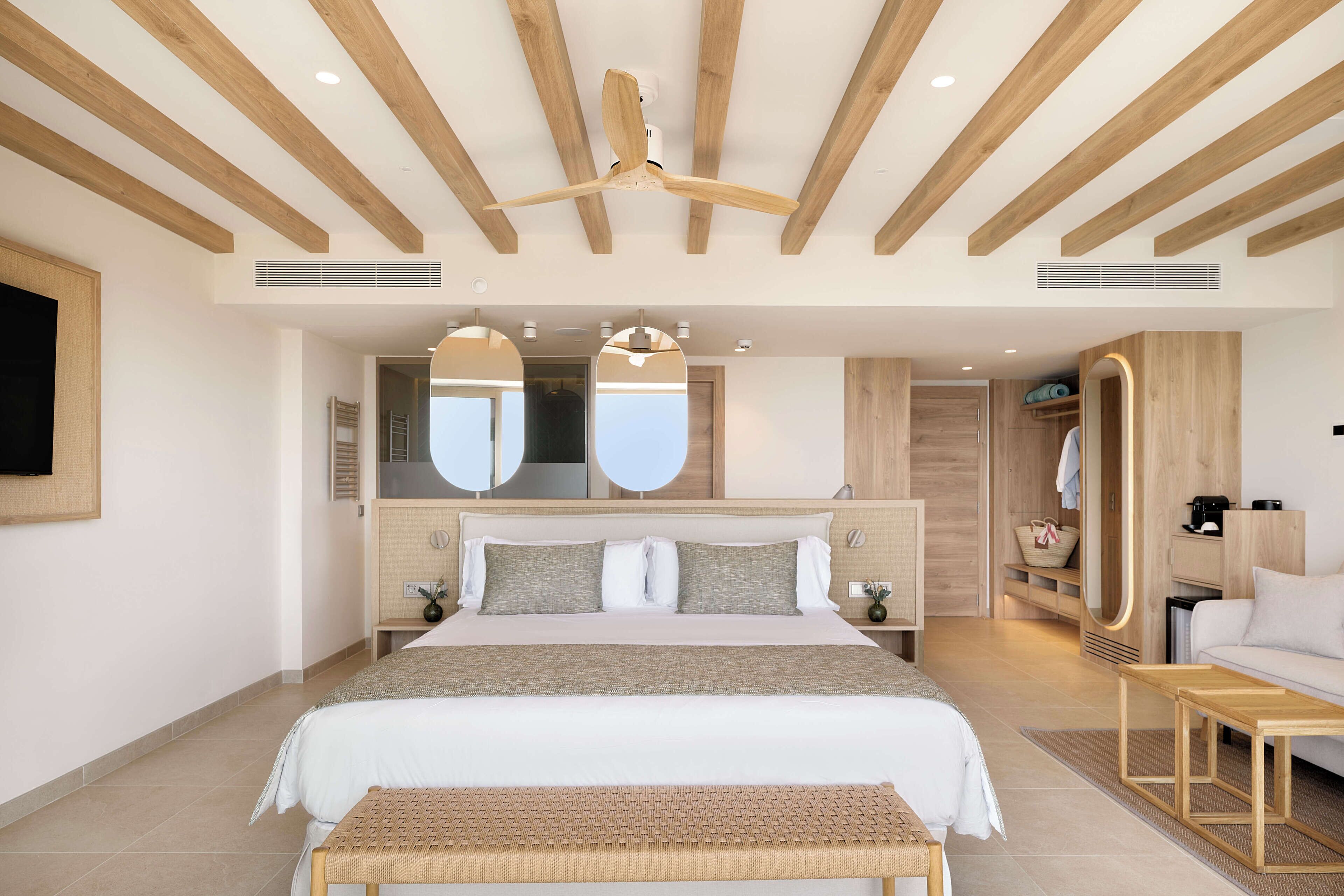 Una habitación de hotel moderna con una cama grande, vigas de madera en el techo, un ventilador de techo, un espejo grande, un sofá y una pequeña mesa.