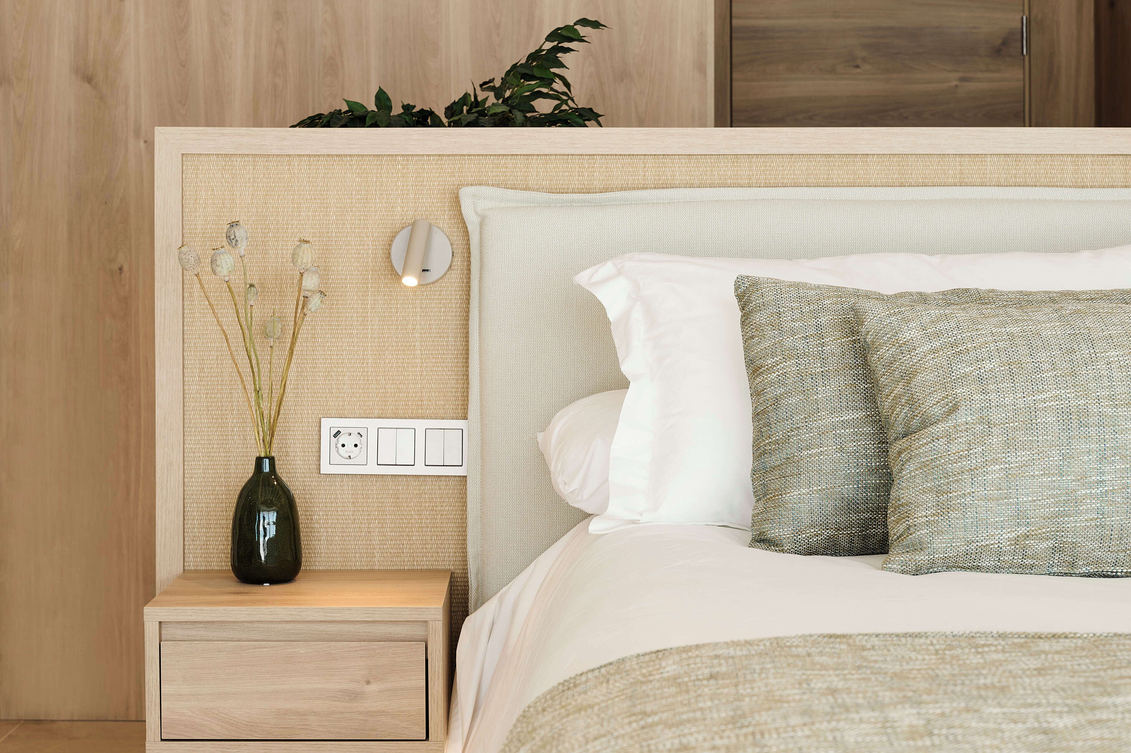A neatly made bed with white and green pillows, a bedside table with a vase of dried flowers, and a wall-mounted light fixture.