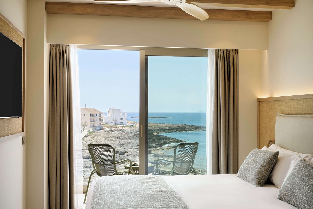 A hotel room with a bed, two chairs, a TV, and a large window with a view of the beach and buildings.
