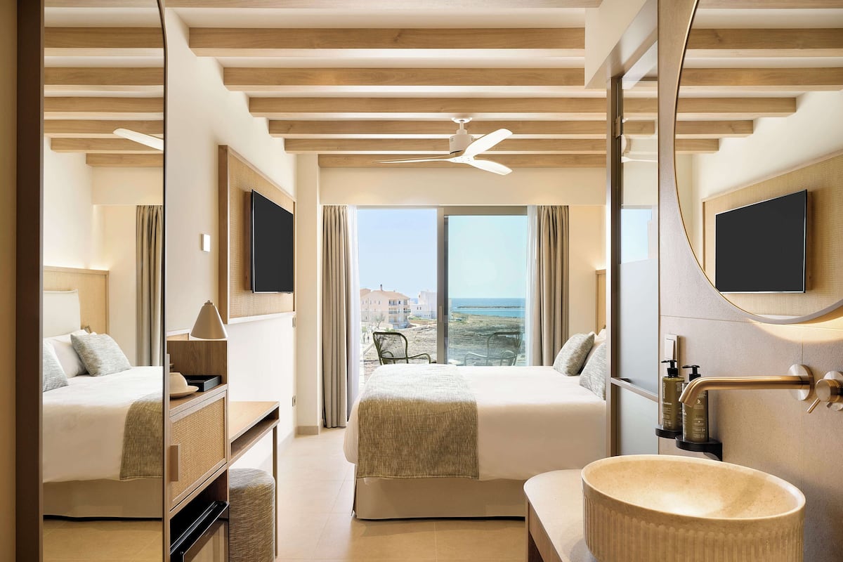 Une chambre d’hôtel avec un grand lit, un coin salon offrant une vue sur la plage, un grand miroir et une salle de bain moderne dotée d’un grand lavabo.