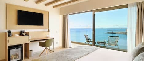 Habitación de hotel con una cama grande, un escritorio con televisor, un refrigerador pequeño y vista al mar.