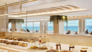 A buffet setup with various food items and condiments, a view of the sea through large windows, and a modern interior design. - Iberostar Selection Es Trenc (Ses Salines)