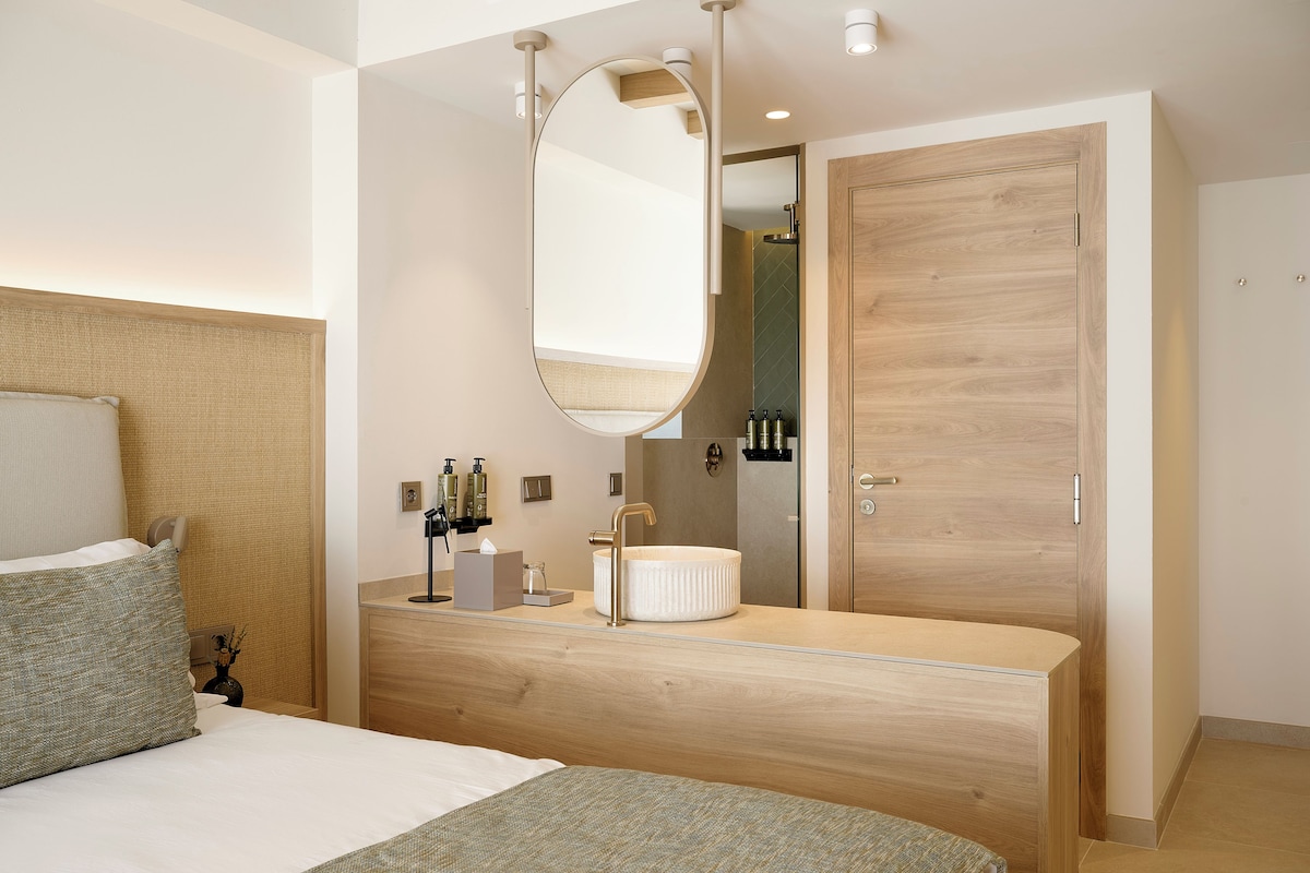 A modern bedroom with a wooden headboard, a round mirror, a bedside table with toiletries, and a wooden wardrobe.