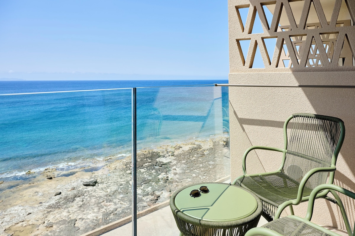 A balcony with a glass railing overlooking a beach and the sea. There are two green wicker chairs and a small round table with a pair of sunglasses on it.