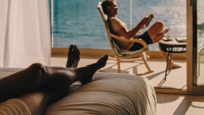 A person lying on a bed with a view of the ocean through a large window. - Iberostar Selection Es Trenc (Ses Salines)