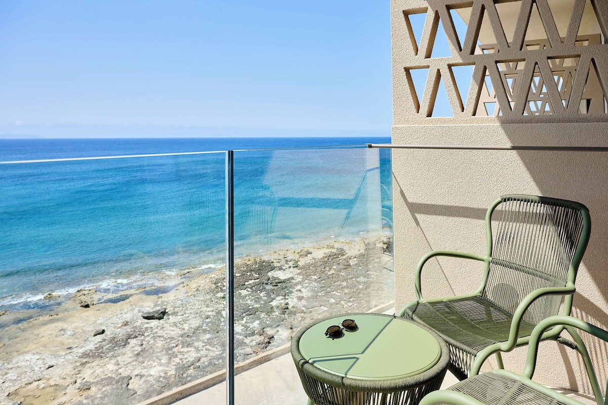 Un balcon doté d’une balustrade en verre, d’où l’on a vue sur une plage et la mer. On y trouve deux fauteuils en osier vert et une petite table ronde sur laquelle sont posées une paire de lunettes de soleil.