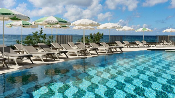 A poolside area with lounge chairs and umbrellas, a clear blue sky, and a view of the sea. - Iberostar Selection Es Trenc (Ses Salines)