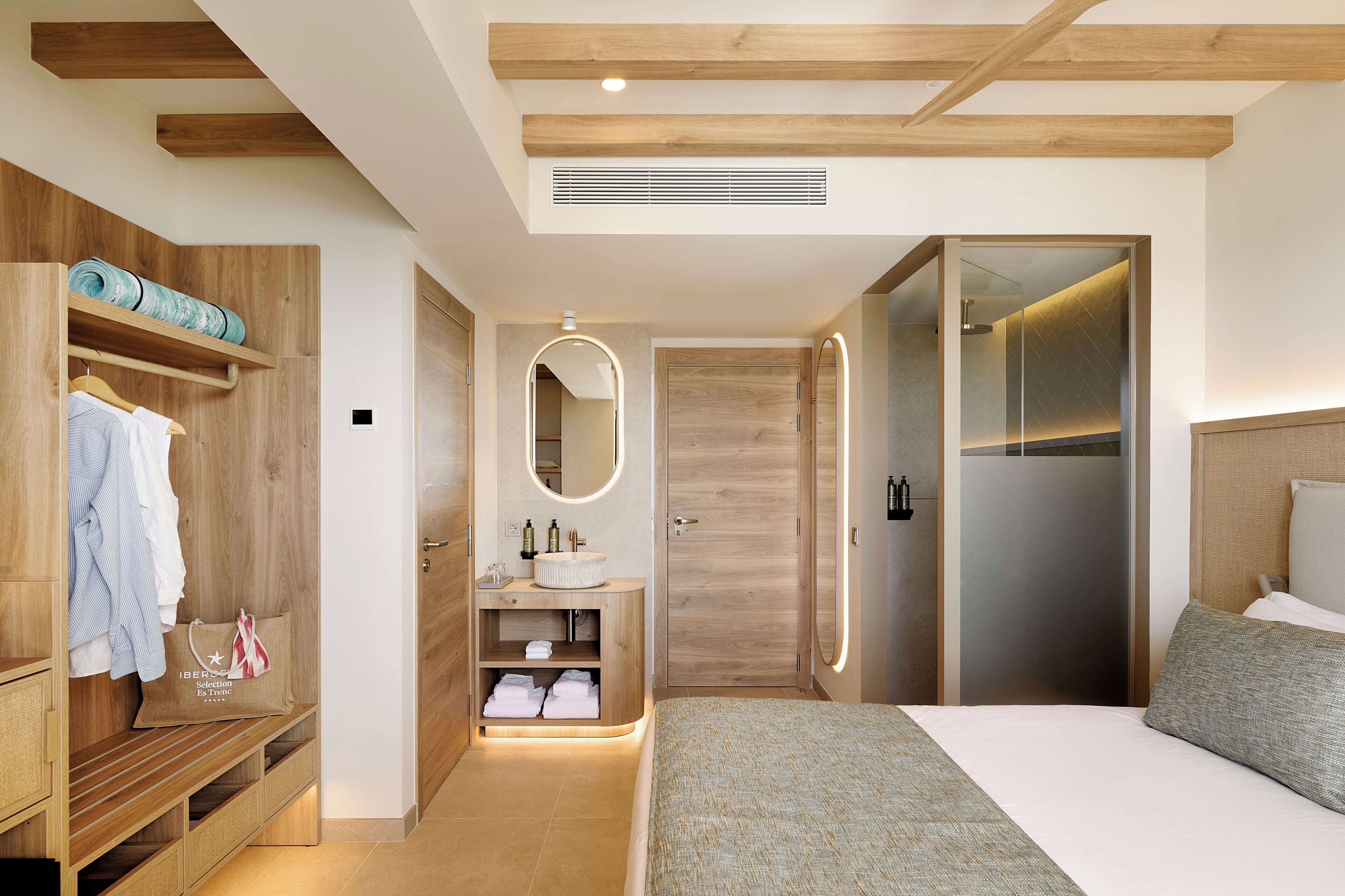 Una habitación de hotel moderna con un armario de madera, una tocador con espejo, una cama con manta gris y un vestidor.
