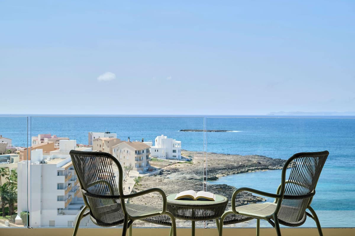 Deux fauteuils en osier, avec un livre ouvert posé sur l’un d’eux, sur un balcon donnant sur une ville côtière et la mer.