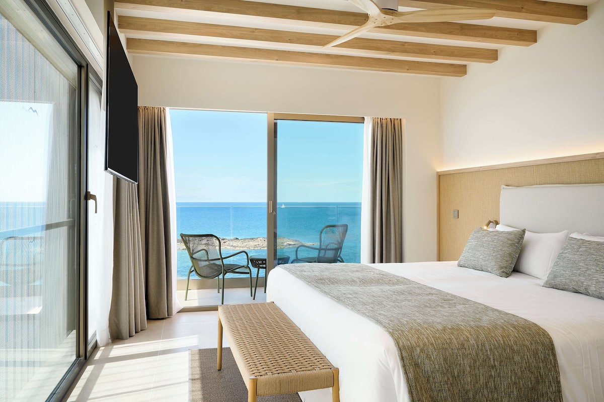 Une chambre d’hôtel avec un grand lit, un banc, une table et une vue sur la mer.