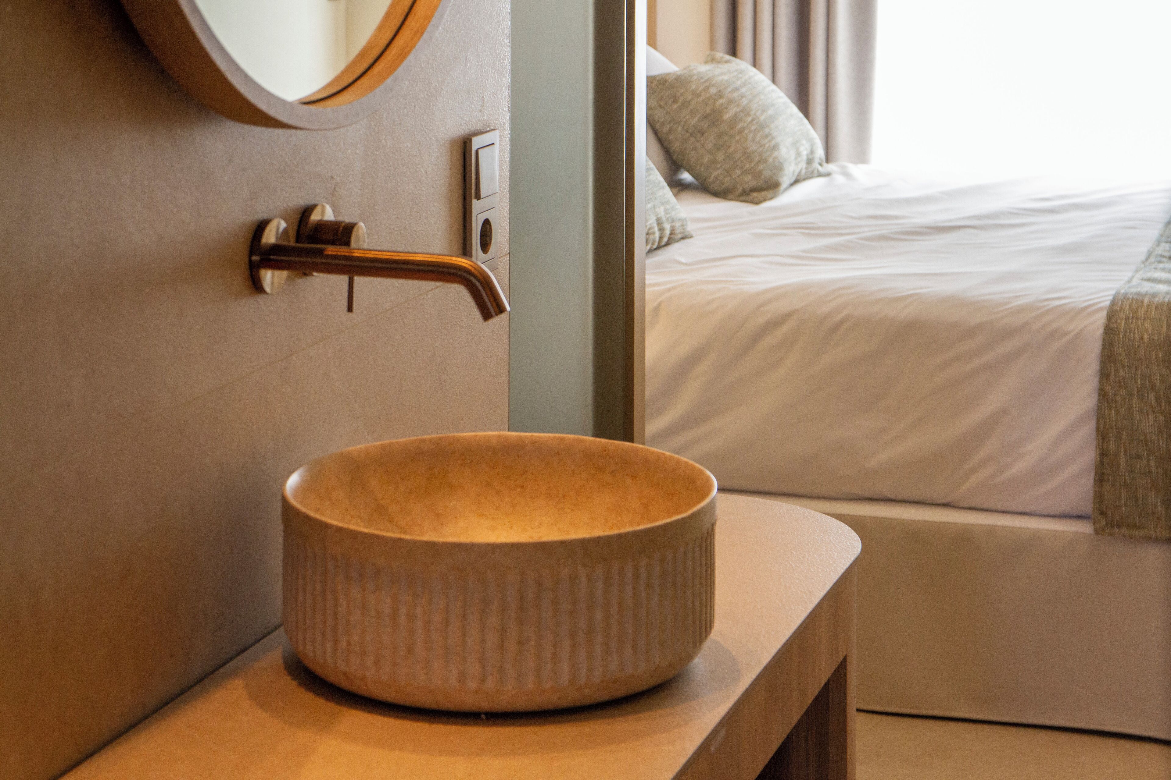 A bathroom with a round ceramic sink, a mirror, and a door leading to a bedroom with a bed and a pillow.
