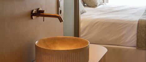 A bathroom with a round ceramic sink, a mirror, and a door leading to a bedroom with a bed and a pillow.