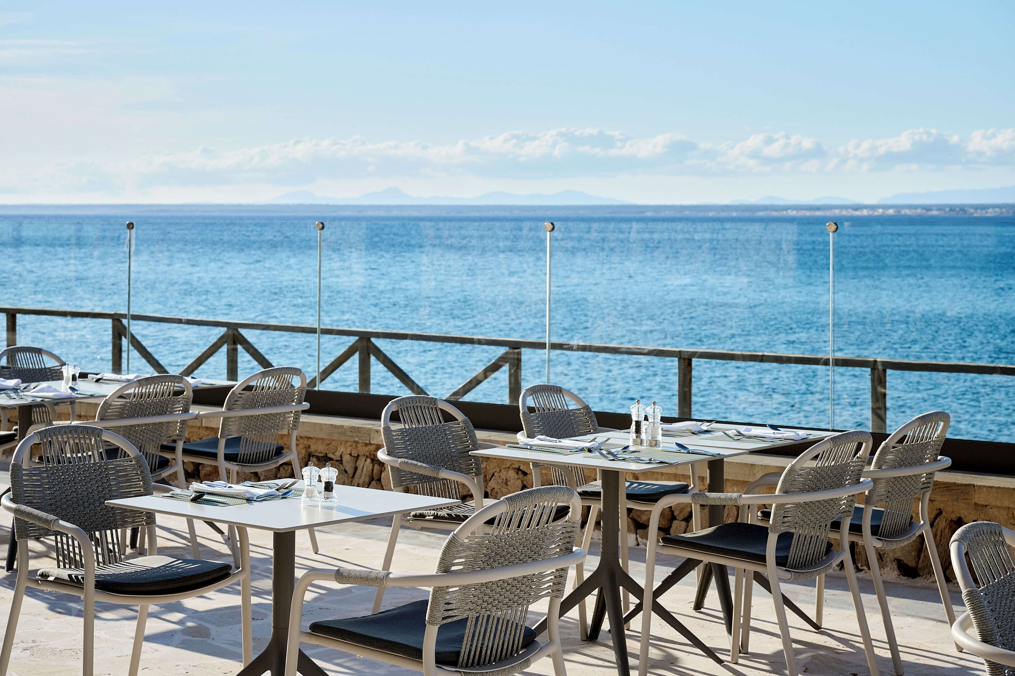 Un espace de restauration en plein air, avec des chaises et des tables en osier, offrant une vue sur une étendue d’eau et sur des montagnes au loin.