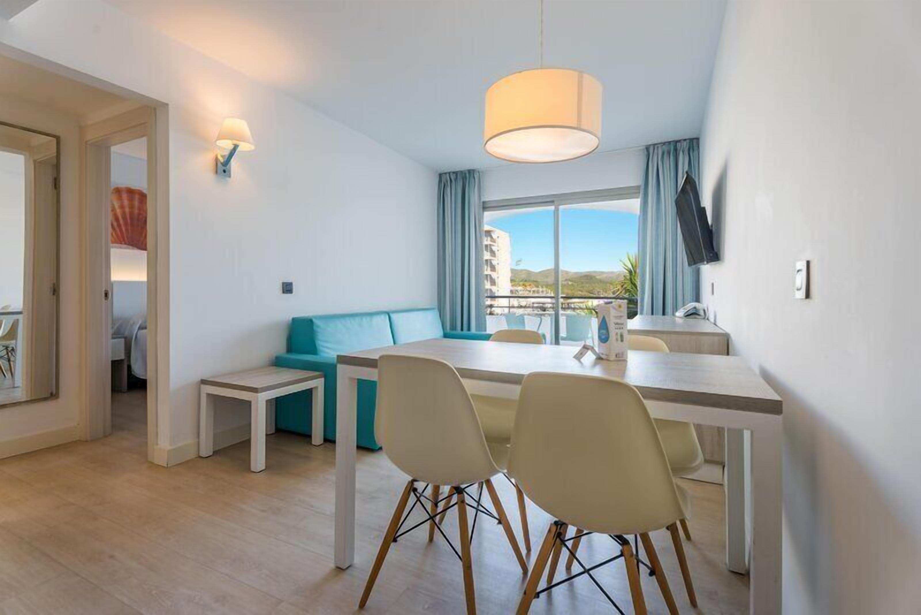 A modern hotel room with a dining area, a sofa, a TV mounted on the wall, and a view of the outside.