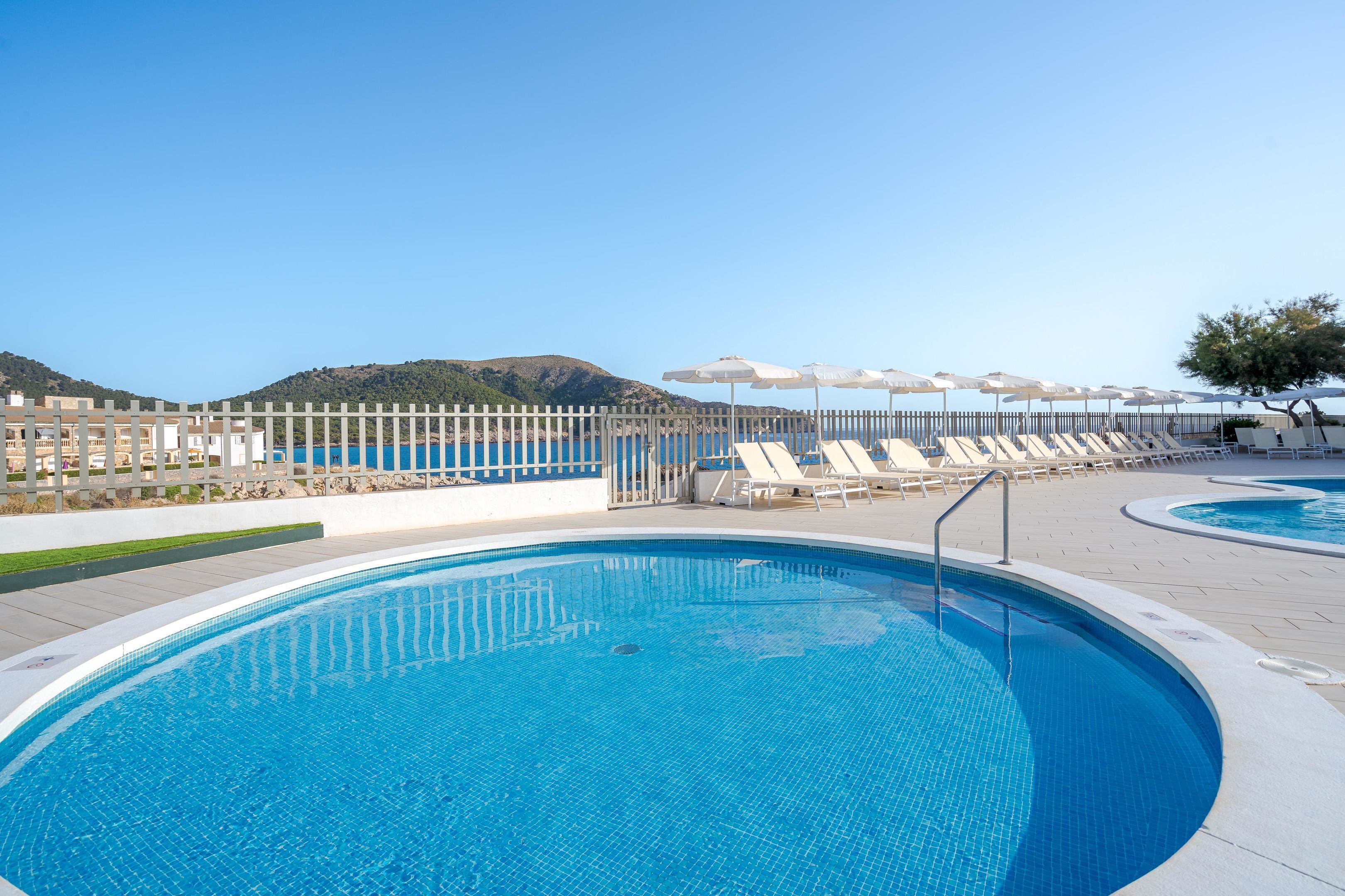 A clear blue pool with lounge chairs and umbrellas, surrounded by a white fence and a view of hills.