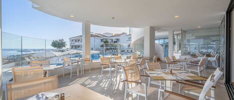 A rooftop dining area with tables and chairs, overlooking a pool and buildings.