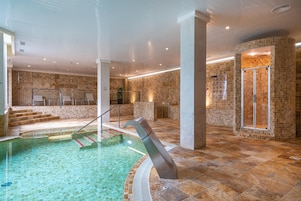 Una piscina interior con agua cristalina, rodeada de paredes con azulejos y una zona de ducha con cerramiento de vidrio.