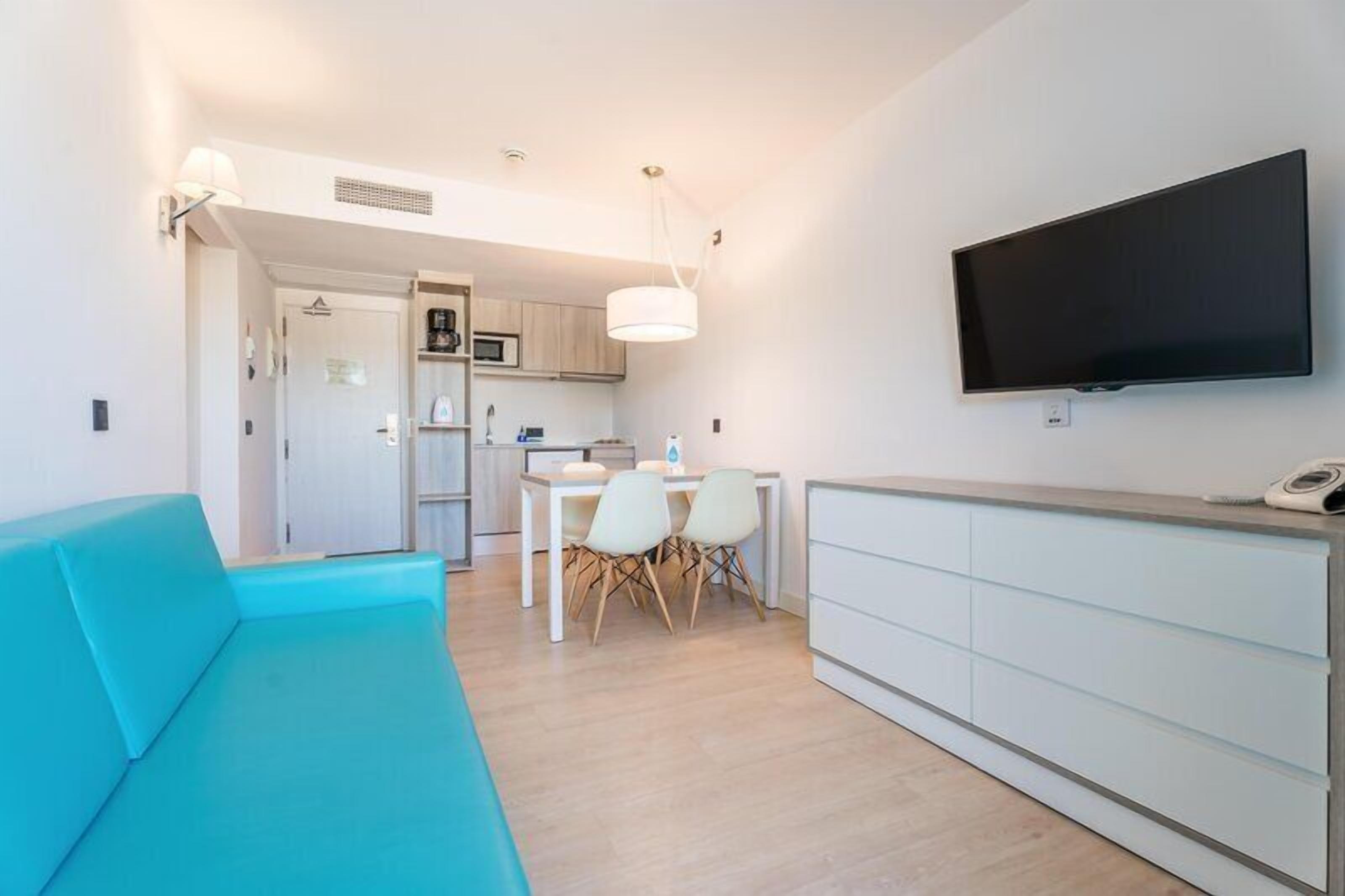A modern living room with a teal sofa, a dining area with a white table and chairs, a kitchen with wooden cabinets, and a mounted TV on the wall.