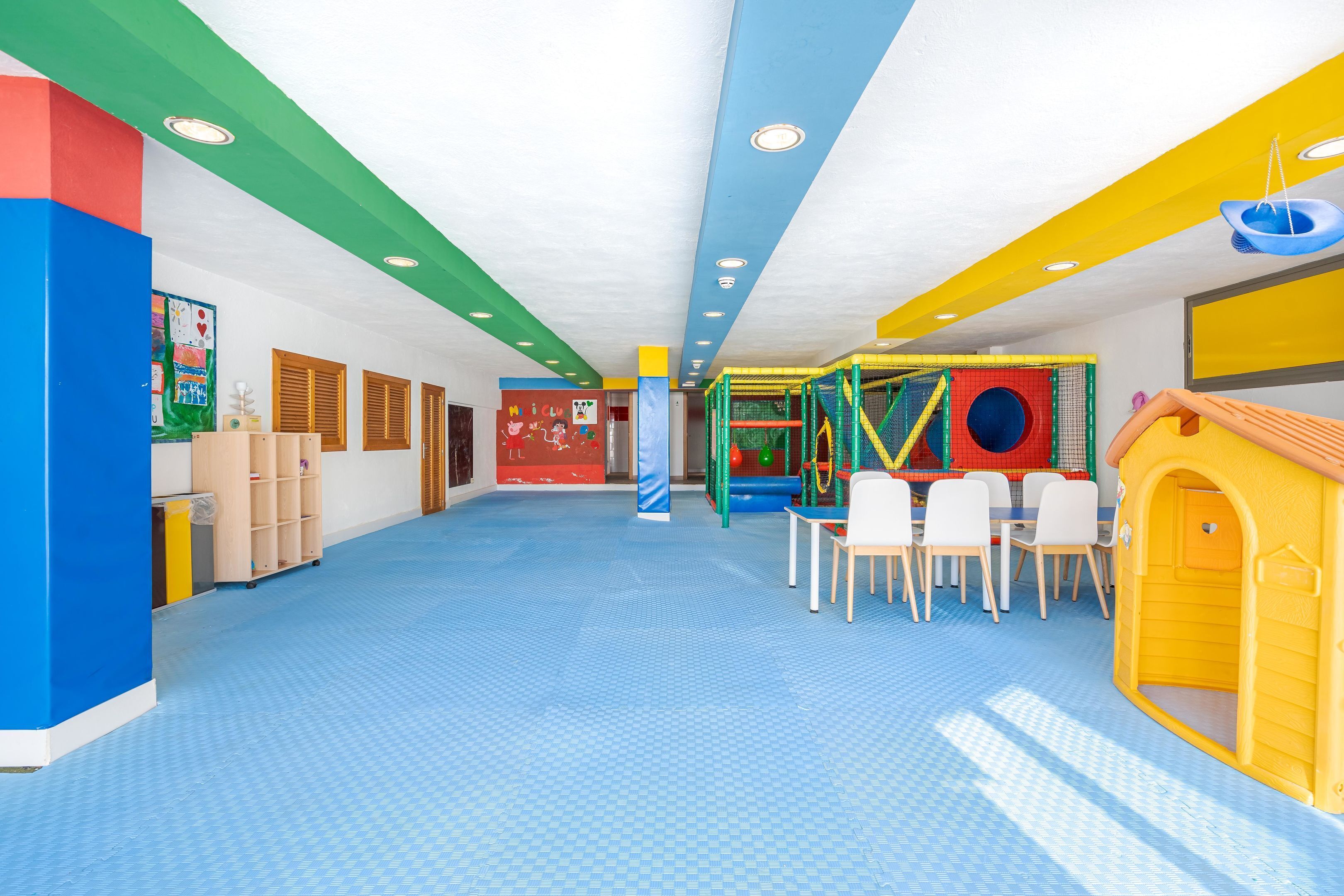 Un área de juegos interior, llena de color, con piso azul, una estructura de juego y una casita amarilla.