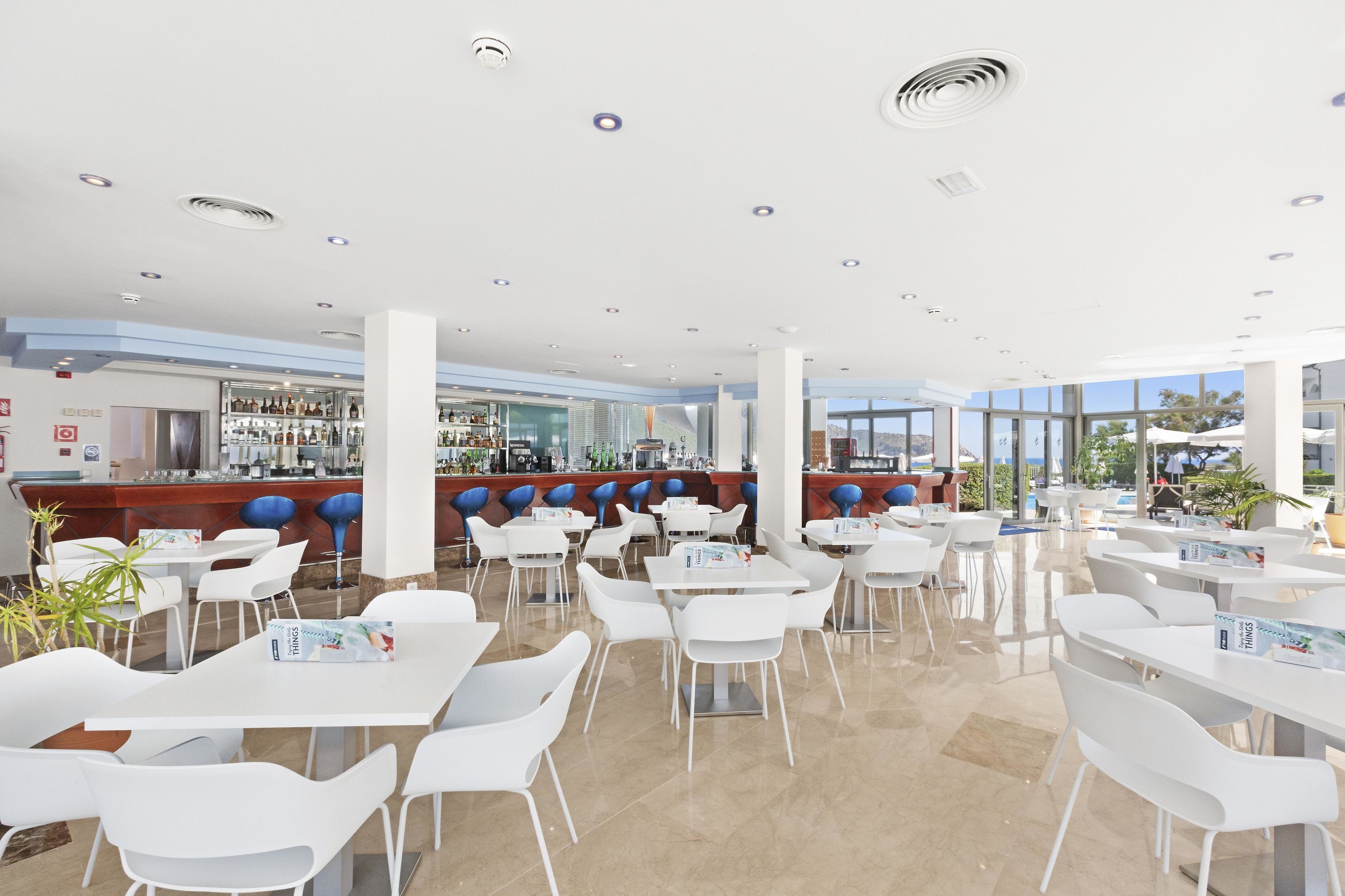 Un espace de restauration intérieur moderne, avec des tables et des chaises blanches, un bar doté de tabourets bleus, et de grandes fenêtres offrant une vue sur l’extérieur.