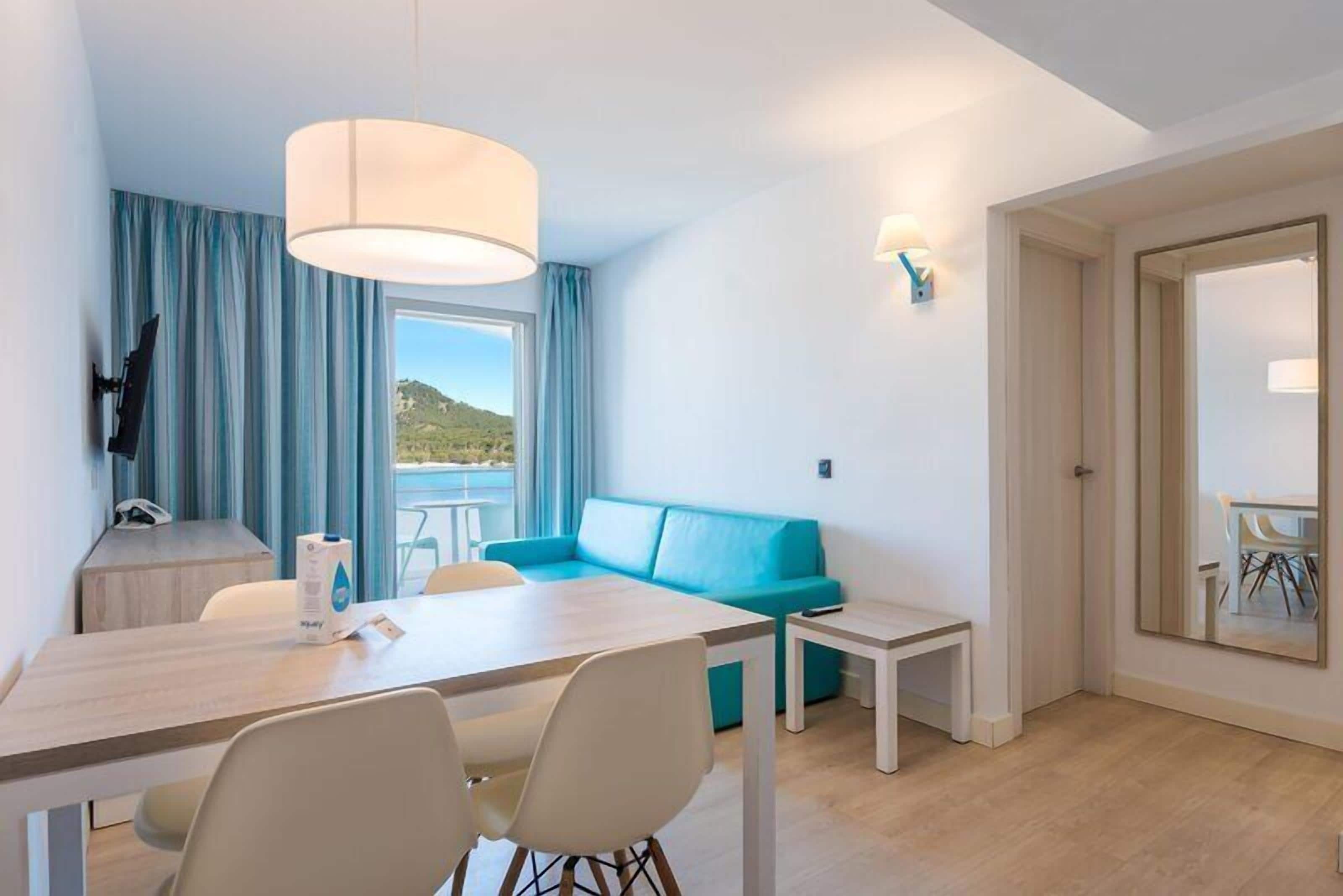 Ein modernes Zimmer mit Esstisch, Stühlen, einer Couch, einem großen Fenster und Blick ins Freie.
