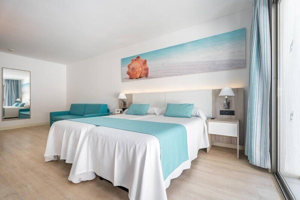 A hotel room with two beds, a large window with curtains, a wall-mounted artwork, and a bedside table with a lamp.