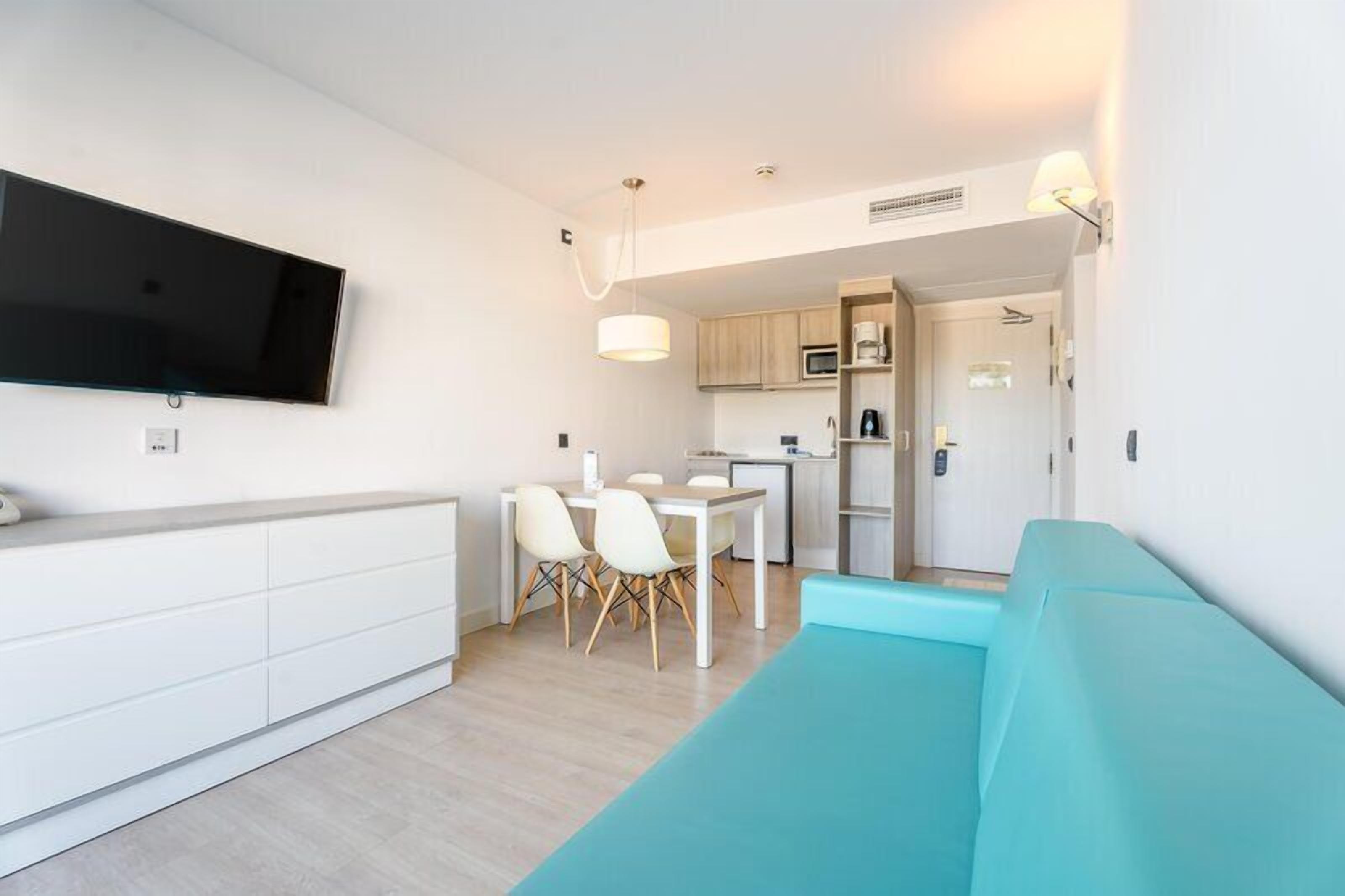 Una sala de estar moderna con un televisor de pantalla plana, un sofá color turquesa, un comedor con sillas blancas y una cocina al fondo.