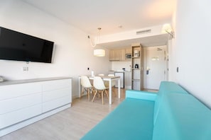 Una sala de estar moderna con un televisor de pantalla plana, un sofá color turquesa, un comedor con sillas blancas y una cocina al fondo.