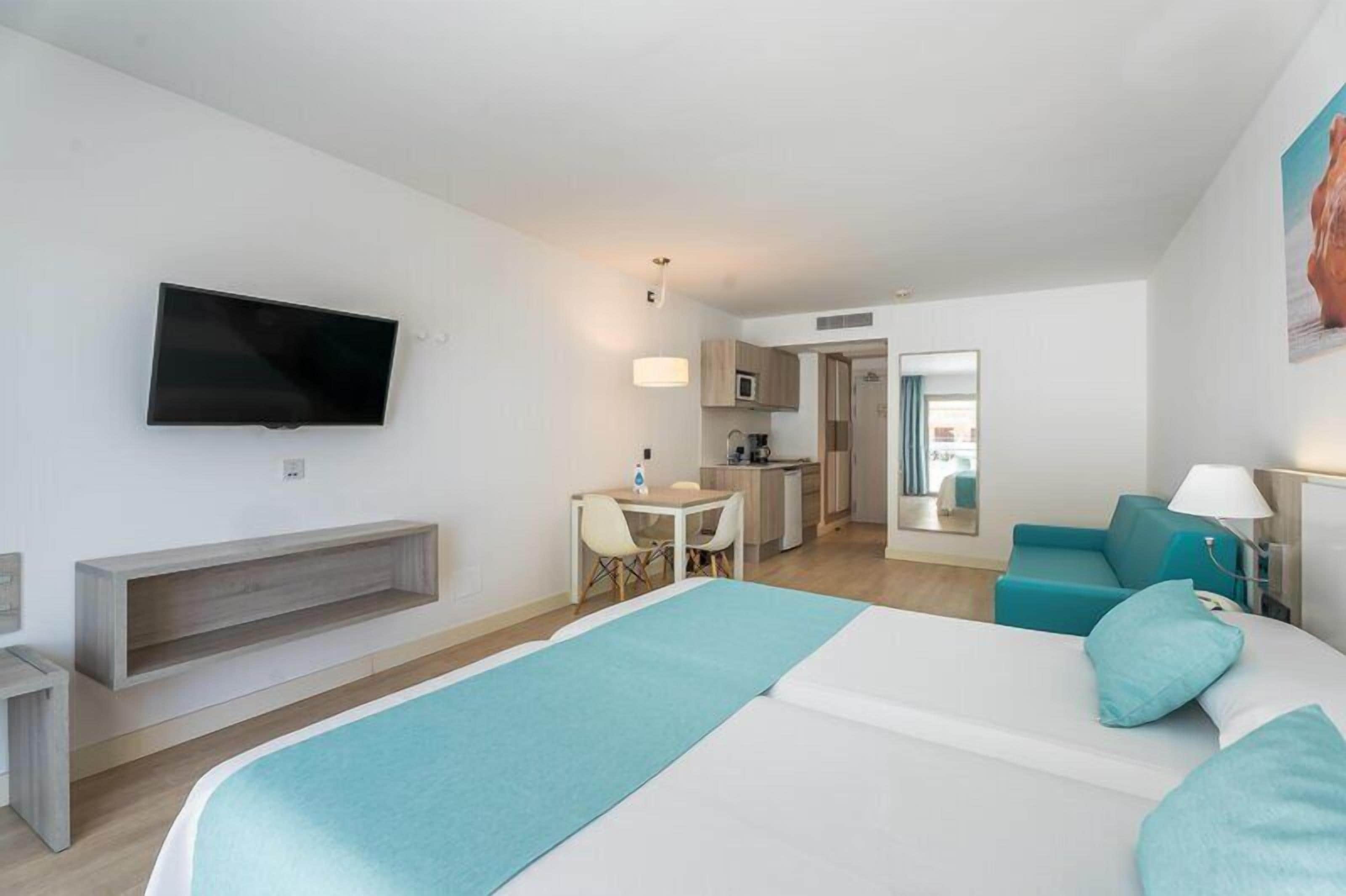 A hotel room with a bed, a TV mounted on the wall, a desk with a chair, a kitchenette, and a bathroom visible through an open door.