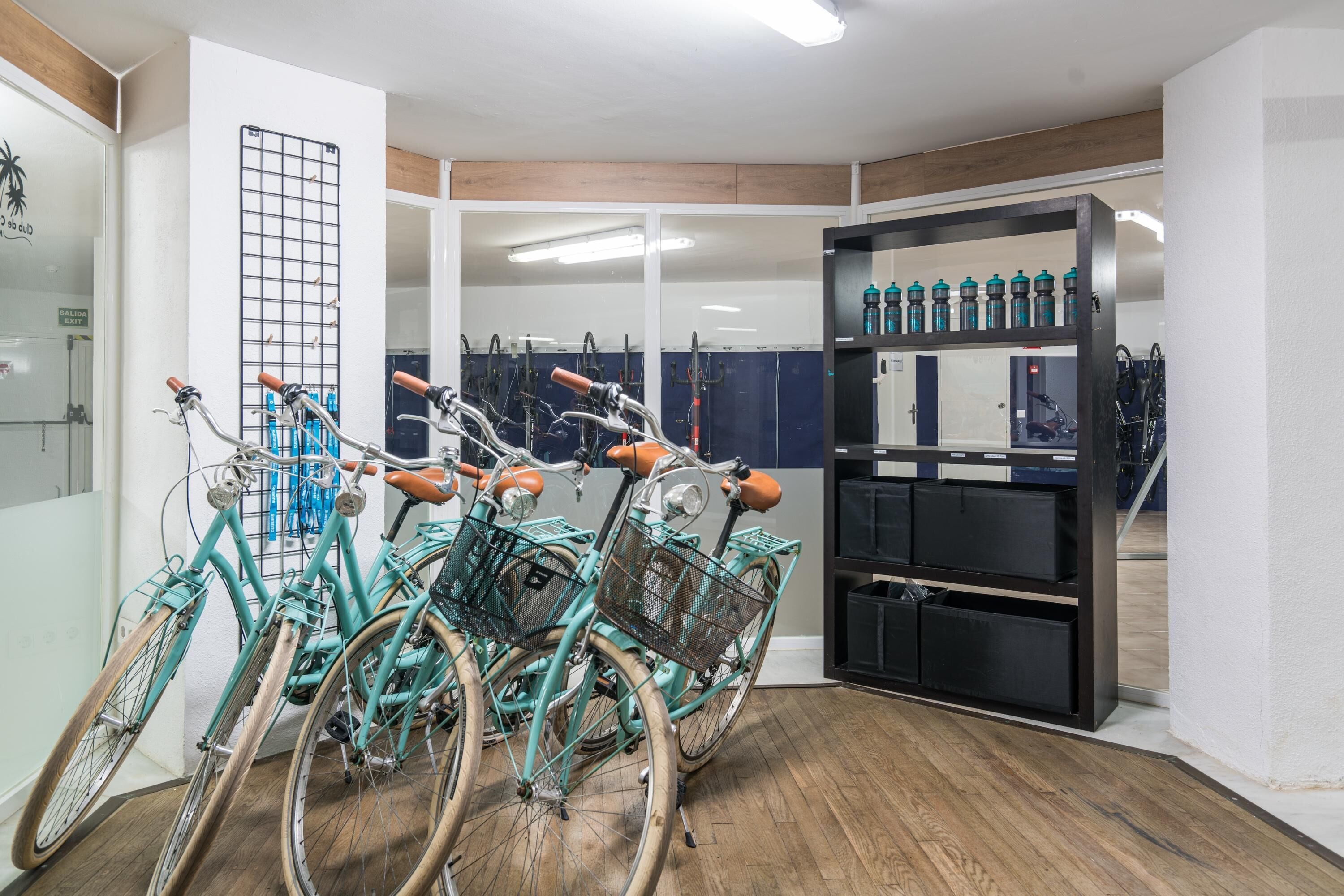 Una habitación con bicicletas estacionadas contra la pared, un soporte con cascos y un estante con botellas de agua.