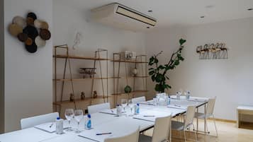 Un salón de conferencias con mesas blancas, sillas y una mesa grande dispuesta para una reunión. Hay estantes con objetos decorativos y una planta grande.