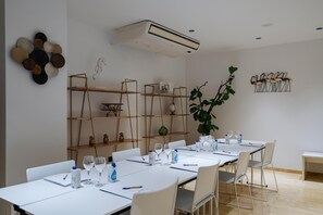 Een vergaderzaal met witte tafels, stoelen en een lange tafel gedekt voor een vergadering. Er staan planken met decoratieve items en een grote plant.